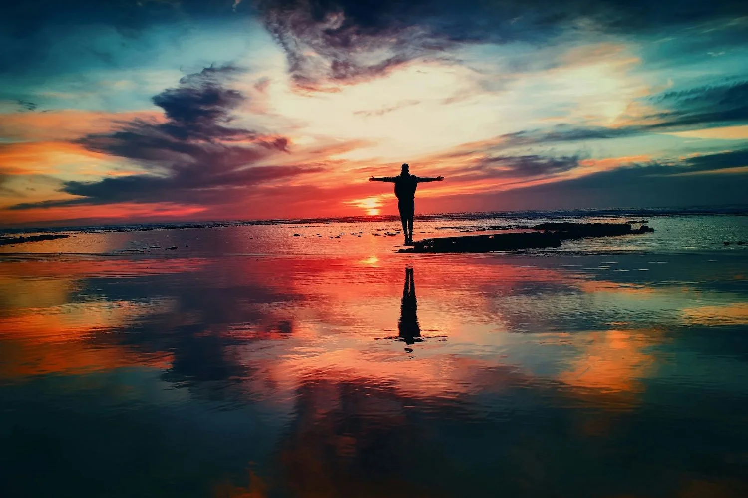 A person standing arms wide at sunrise on a mirror-still shoreline