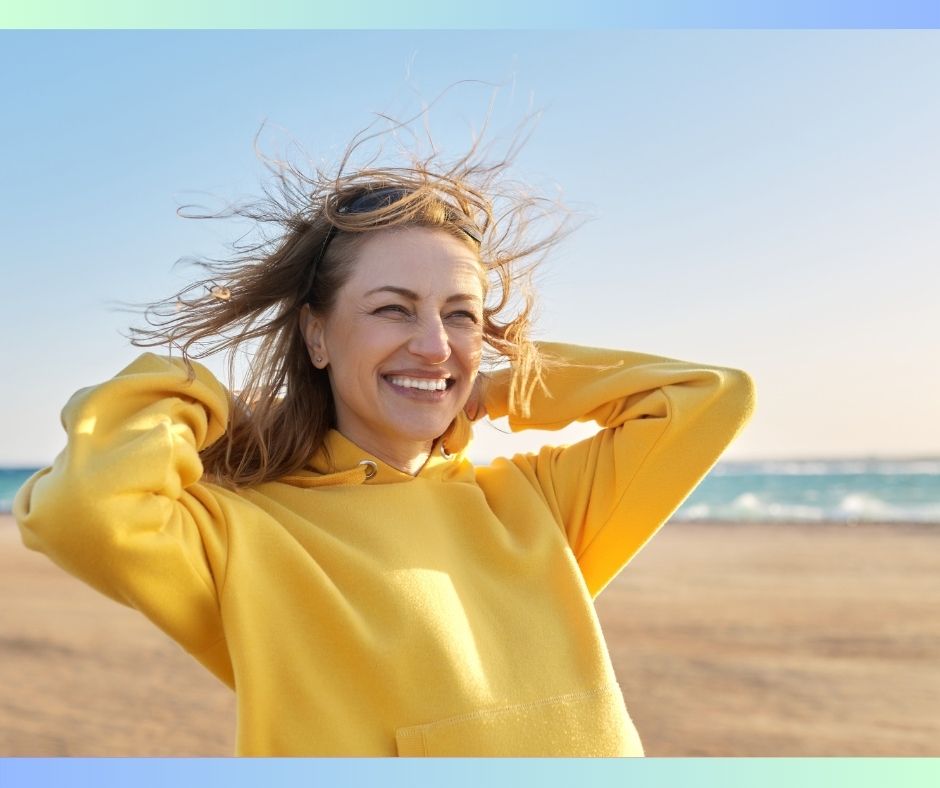 A woman in a yellow fleece hoodie smiling on a sunny beach — living fully and in good health is the goal of personalized cancer screening.